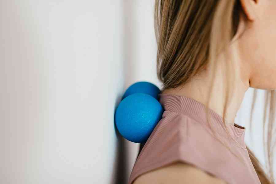 Девушка стоит у стены и массирует шею двумя голубыми массажными шариками